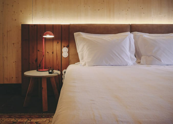Detalhe de uma cama king size do Hotel FeelViana, com o lençol e fronhas brancas, uma cabeceira castanha em camurça e os acabamentos em madeira. 